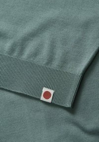 Manche de pull en maille verte avec bord-côtes et petite étiquette blanche comportant un cercle rouge près du bord du poignet.