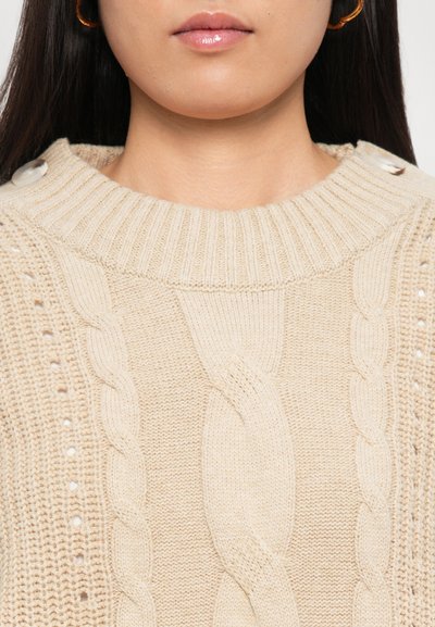 Beige knitted sweater with ribbed collar, cable knit pattern, and button accents on shoulder. Soft texture, mid-weight fabric.