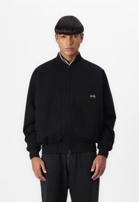 Black bomber jacket with ribbed cuffs and collar. Two large front pockets. Gold logo accent on left chest. Smooth cotton material.