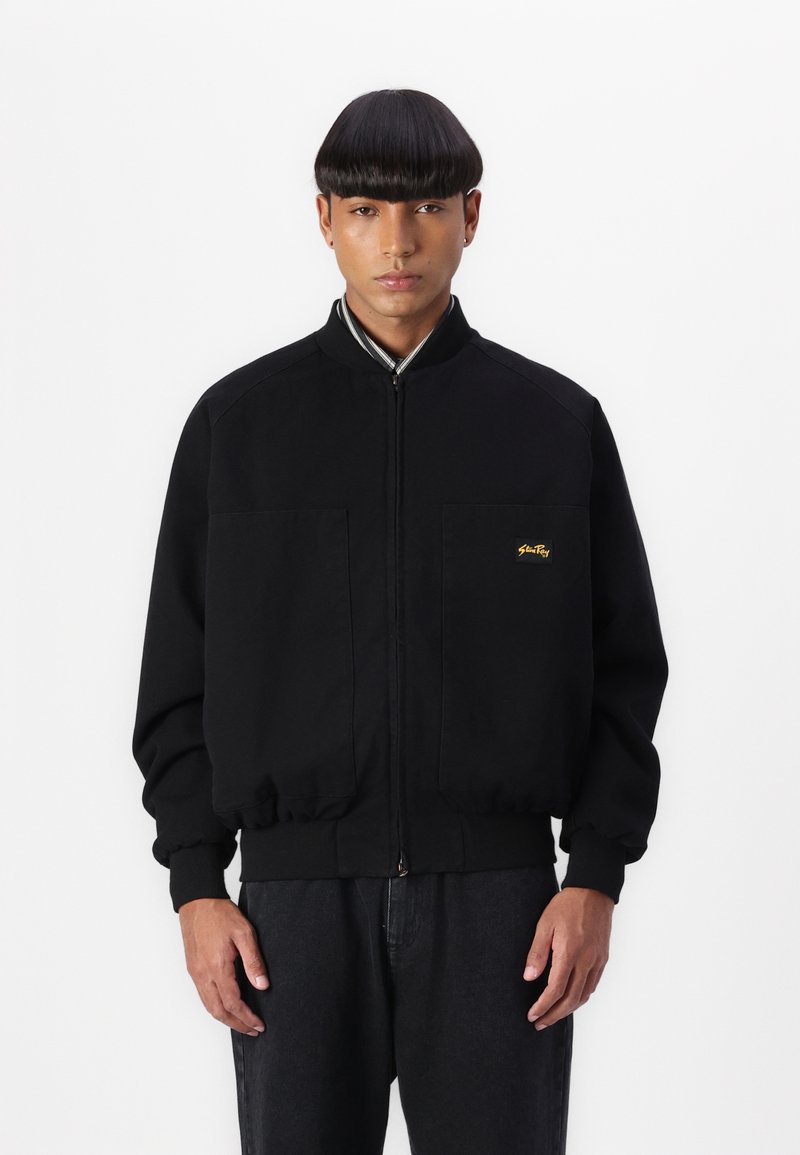 Black bomber jacket with ribbed cuffs and collar. Two large front pockets. Gold logo accent on left chest. Smooth cotton material.