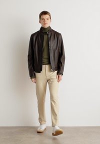 Brown leather bomber jacket, olive turtleneck, beige trousers, white sneakers with gum soles, standing against a neutral background.