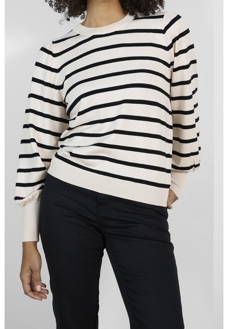 Femme portant un pull crème à rayures horizontales noires et manches bouffantes, associé à un pantalon noir ajusté, se tenant debout avec une main dans la poche.