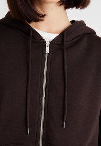 Brown zip-up hoodie featuring a soft texture, drawstring ties, and a silver metal zipper. Layered over a white T-shirt.