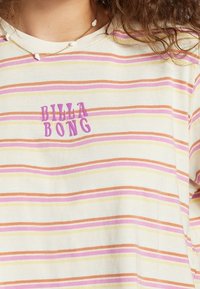 Camiseta de rayas en tonos crema, rosa y naranja con un logo morado de "BILLA BONG". Tela de algodón con un ajuste relajado y cuello redondo.
