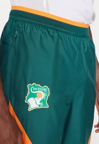 Shorts de sport verts avec des accents orange, une taille élastique et un emblème de la Côte d'Ivoire avec un contour de carte locale et un motif animal.