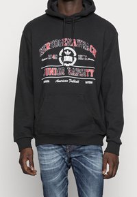 Homme portant un sweat à capuche noir avec le texte rouge et blanc "Herzogenaurach Junior Varsity", le logo Adidas, et un jean bleu sur un fond uni.