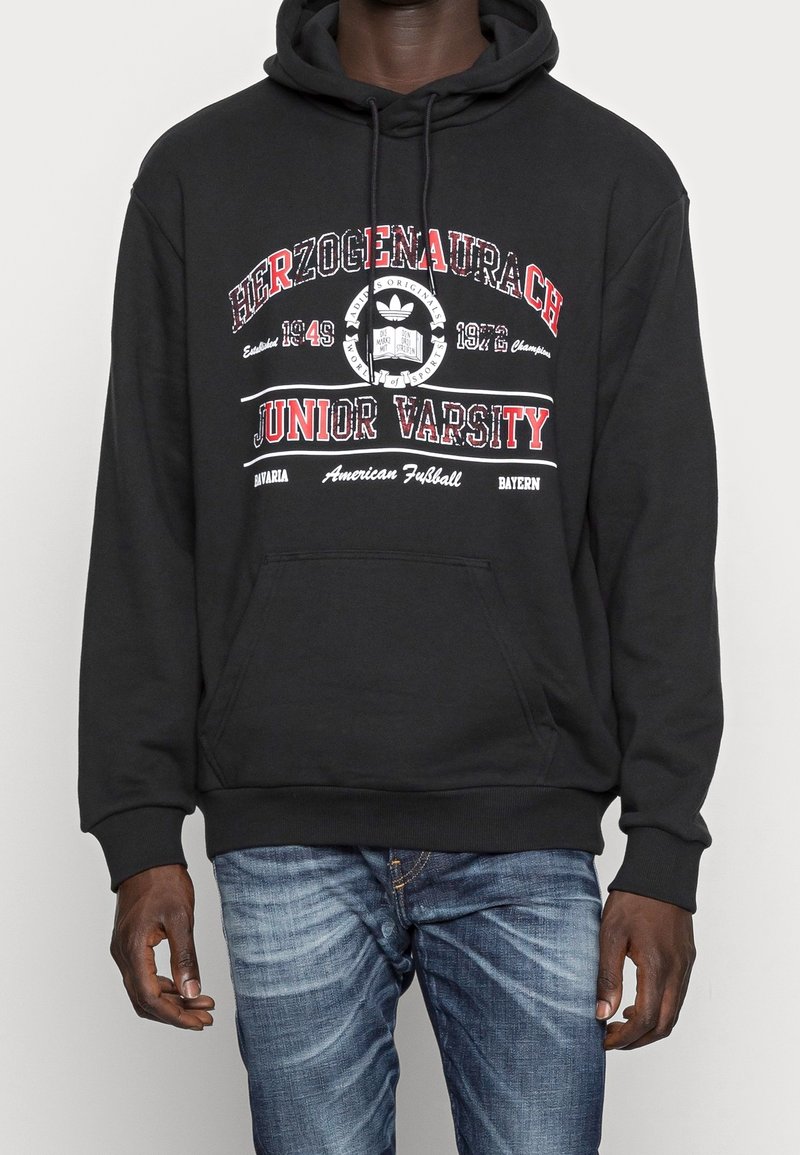 Homme portant un sweat à capuche noir avec le texte rouge et blanc "Herzogenaurach Junior Varsity", le logo Adidas, et un jean bleu sur un fond uni.