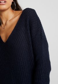 Marinblått stickat tröja med djup V-ringning, texturerad tyg och sänkta axlar. Har en avslappnad passform och ribbat mönster på ärmarna.