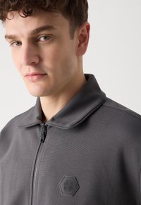 Giacca grigia con zip e colletto alto, in tessuto texturizzato, con un logo esagonale sul petto. Presenta dettagli di cucitura a contrasto.