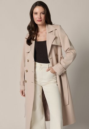 Kobieta o długich ciemnych włosach, ubrana w beżowy trencz na czarnym topie i kremowych spodniach, stoi na tle jednolitego jasnego tła.