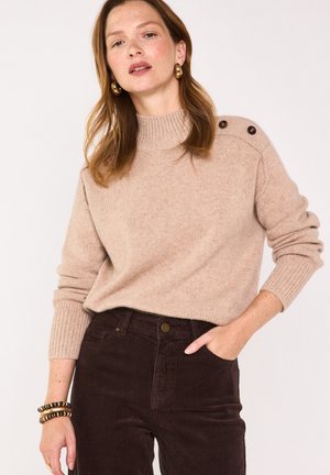 Donna che indossa un maglione beige a maglia con bottoni sulla spalla, pantaloni di velluto a coste marrone scuro, orecchini a cerchio dorati e braccialetti, che posa con la mano in tasca.