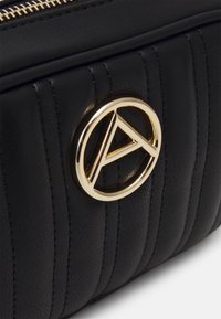 Borsa nera in pelle trapuntata con un logo circolare dorato in evidenza con un design a "A", che mostra motivi testurizzati e una chiusura con zip.