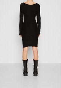 Černé dlouhé rukávy bodycon šaty s výstřihem ve tvaru lodičky, vyrobené z měkké tkaniny. Kombinováno s vysokými černými kozačkami na podpatku.