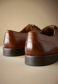 Zapatos de cuero marrón con un acabado suave, tacones bajos de goma negra y un diseño clásico de cordones, que muestran un detallado acabado en las costuras y la forma.