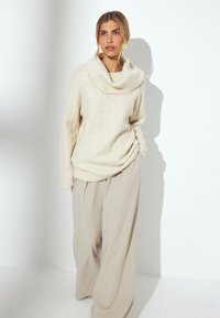 Maglione lavorato a maglia color crema con collo a lupetto, caratterizzato da un motivo a diamante testurizzato. Abbinato a pantaloni beige a gamba larga. Illuminazione brillante.