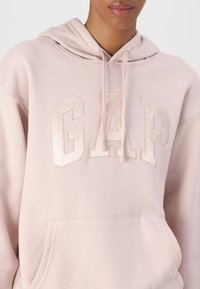 Różowa bluza z kapturem wykonana z miękkiego materiału, z dużym, błyszczącym logo "GAP". Posiada przednią kieszeń oraz sznurki w kapturze.