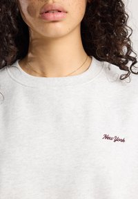 Close-up of a person with curly hair wearing a light gray sweatshirt with "New York" embroidered in burgundy on the chest.