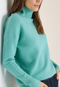 Jasnoturkusowy dzianinowy sweter z golfem o fakturowanym wzorze, z prążkowanymi mankietami i luźnym krojem, w połączeniu z ciemnymi dżinsami.