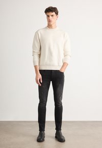 Suéter de canalé crema con escote redondo, jeans ajustados negros y botines negros, de pie sobre un suelo de color neutro.