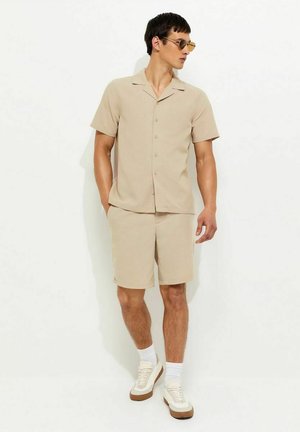 Mężczyzna ubrany w beżową koszulę z krótkim rękawem zapinaną na guziki i pasujące krótkie spodenki, białe skarpetki, białe trampki i brązowe okulary przeciwsłoneczne, stojący na białym tle.