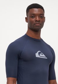 Sötétkék, rövid ujjú rash guard, sima, rugalmas anyagból készült, fehér logóval a mellrészen, raglán ujjal.