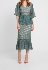 Spetsklänning med peplum-topp i teal, med V-ringning, utsvängda ärmar och ett mönstrat kjol med volangkant. Utrustad med en figurnära midja.