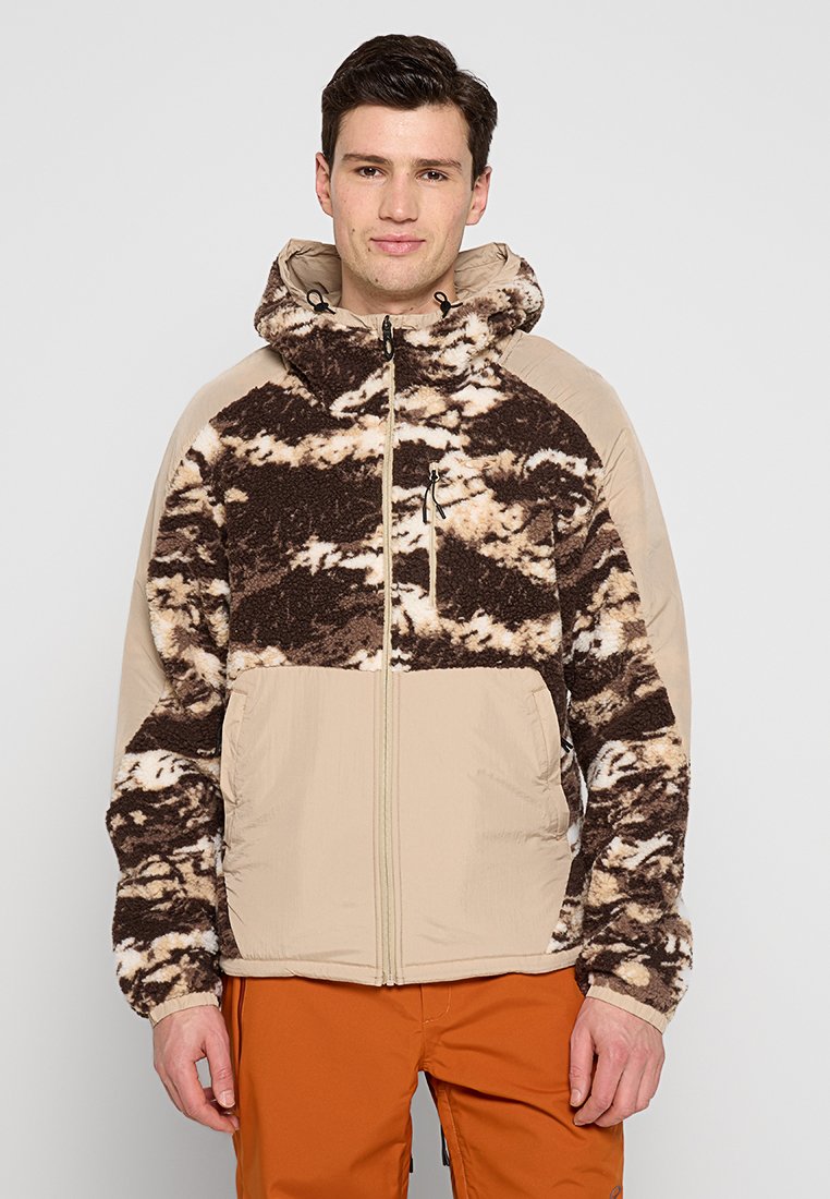 Veste en polaire marron et crème avec capuche, présentant une partie supérieure à motifs et une partie inférieure unie ; poches zippées à l'avant.
