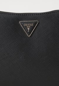 Borsa a tracolla in tessuto nero con texture, caratterizzata da un logo metallico triangolare dorato con la scritta "GUESS" e "USA". Chiusura a zip nella parte superiore.
