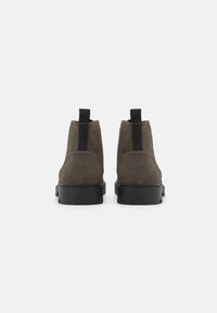 Botines de ante verde oliva oscuro con una textura suave, que cuentan con suelas de goma negras y lengüetas en la parte trasera para facilitar el uso.