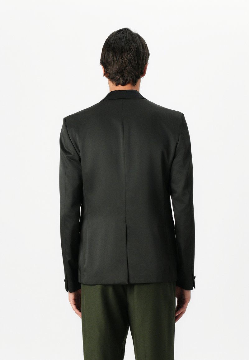 Blazer noir ajusté avec revers crantés, coupe standard, tissu lisse, fente simple au dos, porté sur un pantalon vert.