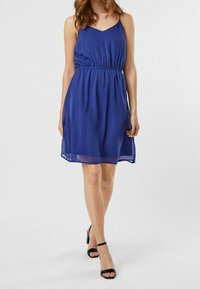 Blå ärmlös klänning med v-ringning och resår i midjan, som faller ovanför knät. Transparent tyg med slät yta. Svarta sandaler med klack.