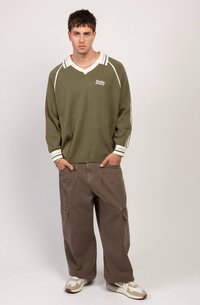 Νεαρός άνδρας που στέκεται με τα χέρια στις τσέπες, φορώντας ελαιόλευκο πουλόβερ, φαρδιά καφέ cargo παντελόνια και μπεζ αθλητικά παπούτσια σε απλό φόντο.