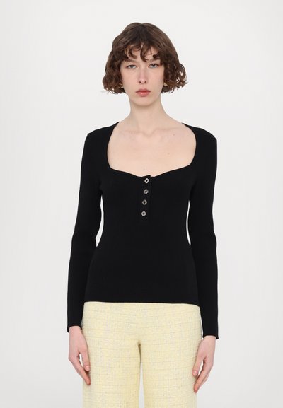 Pull en maille noir avec un décolleté carré, des manches longues et une patte de boutonnage comportant cinq boutons métalliques. Porté avec un pantalon de couleur claire.