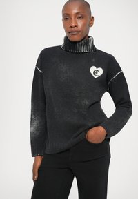 Suéter preto de malha com gola alta, apresentando um design de coração, decorado com um detalhe bordado em branco e uma textura de gradiente subtil perto da bainha.