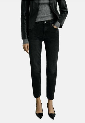 Frau trägt schwarze Jeans mit hoher Taille, grauen gerippten Pullover, schwarze Lederjacke und schwarze spitze High Heels, steht mit einer Hand in der Tasche.