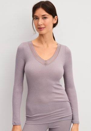 Frau mit dunklen Haaren, die ein hellviolettes geripptes Langarmshirt mit Spitzenbesatz am V-Ausschnitt trägt, steht vor einem einfarbigen hellen Hintergrund.