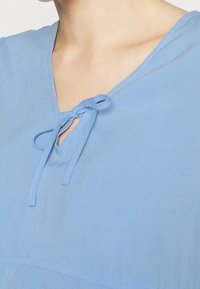 Světle modrý top s výstřihem do V a detailem v podobě šňůrky na výstřihu. Hladká textura a uvolněný střih s jemnými švy.