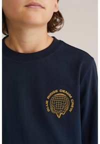 Asmuo, vilkintis tamsiai mėlyną džemperį su apvalia iškirpte, ant kurio pavaizduotas geltonas pasaulio dizainas ir tekstas "Blue Ridge Denim Crew" ant krūtinės.