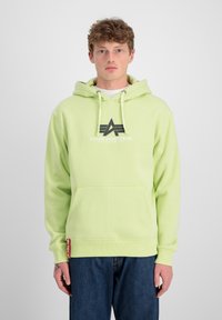 Sudadera verde claro con cordones y bolsillo frontal. Presenta una impresión de logo en negro. Combinada con vaqueros azules. Textura suave.