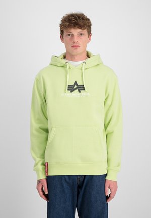 Sudadera verde claro con cordones y bolsillo frontal. Presenta una impresión de logo en negro. Combinada con vaqueros azules. Textura suave.