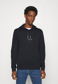 Sudadera negra con capucha ajustable, fabricada en tejido suave, y un pequeño logo en el pecho. Combinada con jeans azules, llevada por un modelo frente a un fondo neutro.