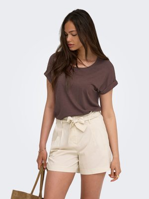 Frau mit langen dunklen Haaren, die ein braunes kurzärmeliges Oberteil und beige hoch taillierte Shorts mit einer Schleife trägt, hält eine braune Tragetasche.