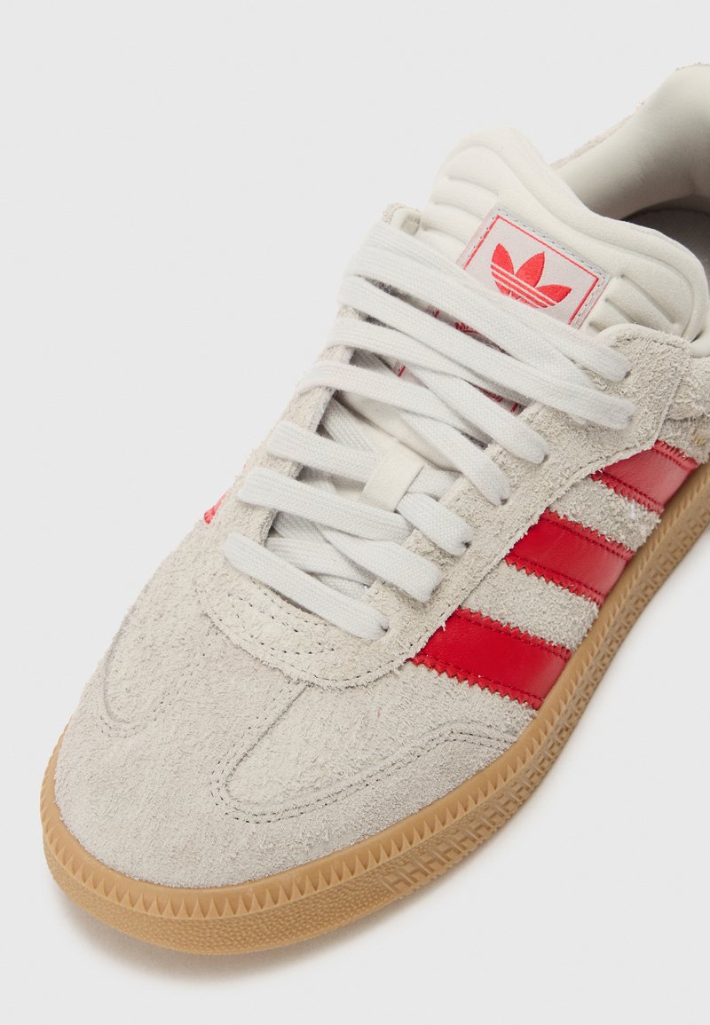 Baskets en suédine blanche avec des rayures en cuir rouge et une semelle en caoutchouc gomme. Elles présentent des lacets blancs et une languette rembourrée avec un accent de logo.