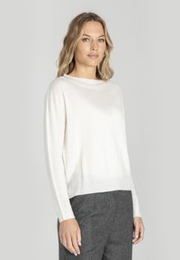 Kobieta o włosach blondu do ramion, ubrana w długi biały sweter i szare spodnie, stojąca na jednolitym tle.