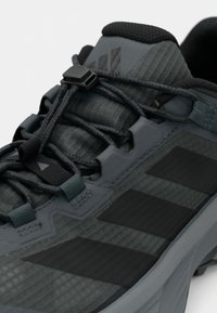 Σκούρο γκρι αθλητικό παπούτσι με υφή στην επάνω επιφάνεια, μαύρες λωρίδες διακόσμησης και ένα μίνιμαλ σύστημα κορδονιών με ασφαλές κλείδωμα.