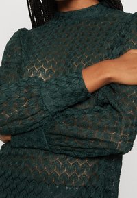 Grön texturerad blus med ett dekorativt mönster, som har en hög halsringning och långa ärmar. Tyget verkar lätt och intrikat vävt.