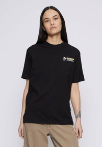 Noor, kellel on pikad tumehigised juuksed, kannab musta graafilise t-särki ja beeže pükse, seistes heledal ühtlasel taustal.