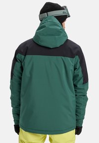 Grün-schwarze Jacke mit Kapuze, aus glattem Material. Verfügt über einen Reißverschluss und mehrlagige Farben auf den Schultern.