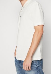 Polo blanco de manga corta con cuello acanalado, que presenta tres botones y un pequeño logo. Combinado con unos jeans de mezclilla azul claro.