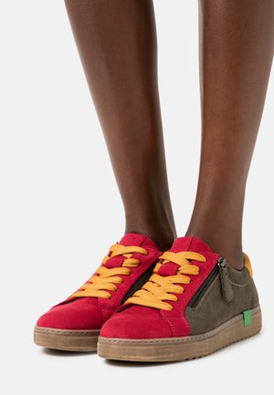 Jambes à peau foncée portant des baskets basses avec l'avant rouge, les côtés olive, des lacets jaunes, une fermeture éclair latérale et des semelles marron clair sur un fond uni.